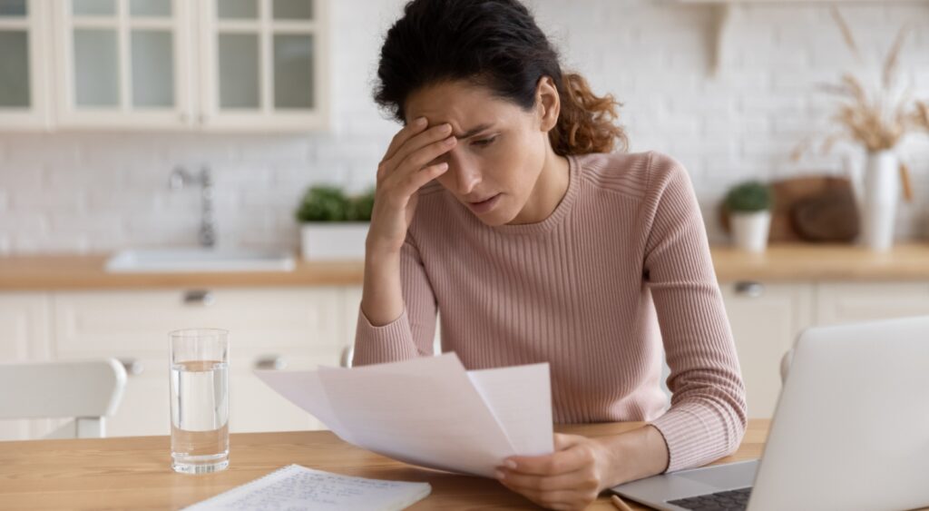 Stressed young woman looking at paperwork while using laptop.