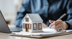 A person signing a document with a model house on the table.